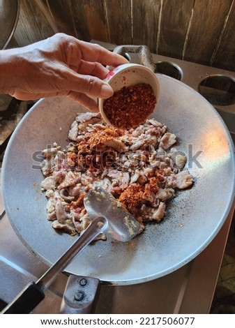 The cooking picture shows several pork slices in a silver pan with the hands of an elderly woman pouring finely chopped dried chili from a cup onto the pork in a pan with a black spatula overturned.