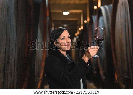 Woman in the wine cellar with barrels in background drinking and tasting wine. Royalty-Free Stock Photo #2217530319