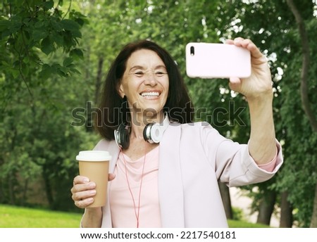 Beautiful elderly brunette woman holding coffee to go and smartphone. Takes a selfie.