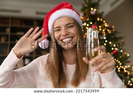 Happy pretty young woman in festive mood holding glass of wine in hands, waving hand looking at camera, recording video or holding web camera call, congratulating with winter holidays Christmas eve. Royalty-Free Stock Photo #2217543999