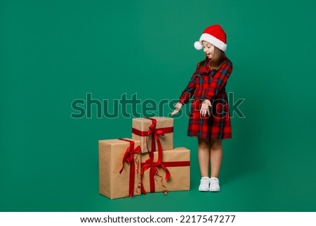 Full body happy little child kid girl 7 year old wear red dress Christmas hat posing look at near present boxes with gift ribbon bow isolated on plain dark green background Happy New Year 2023 concept