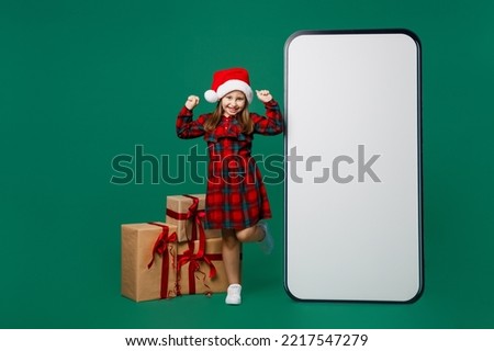 Full body little child kid girl 7 years old wear red dress Christmas hat posing big blank screen mobile phone with area do winner gesture isolated on plain green background Happy New Year 2023 concept