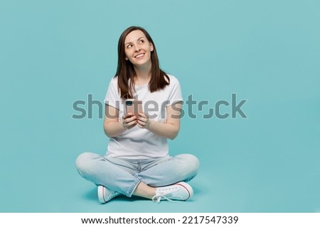Full body young minded fun woman 20s she wear white t-shirt sitting on floor hold in hand use mobile cell phone look aside overhead isolated on plain pastel light blue cyan background studio portrait