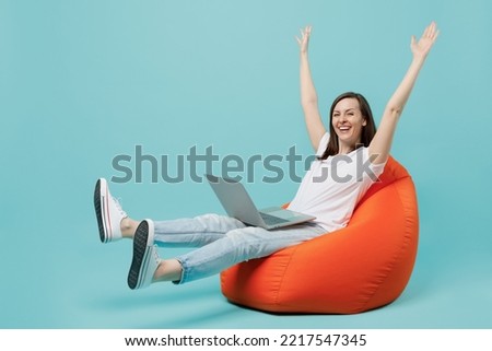 Full body young excited woman 20s she wear white t-shirt sit in bag chair hold use work on laptop pc computer raise up hands finish job celebrate isolated on plain pastel light blue cyan background