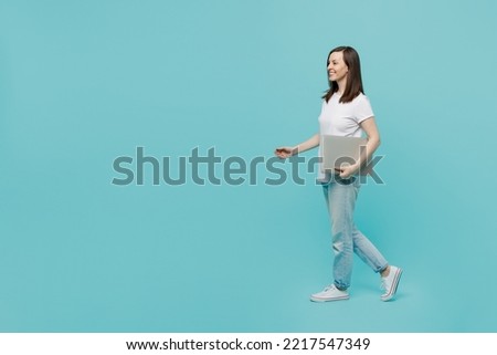 Full body side view young smiling freelancer cool happy fun caucasian woman 20s she wear white t-shirt walking going hold closed laptop pc computer isolated on plain pastel light blue cyan background