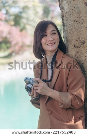 Vertical Asian Women with vintage film camera take a photo. Smiling female photographer look at photo from professional camera outdoor. Vertical photo of young woman shoot photo in green nature park