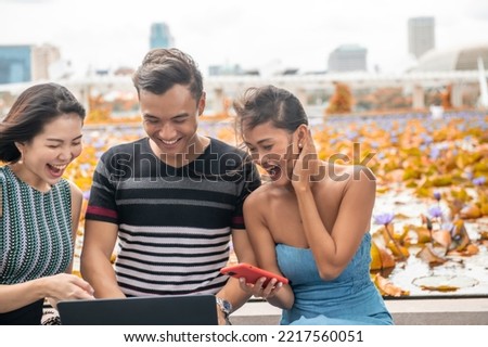 Three happy young asian friends outdoor seated on a public bench using laptop.