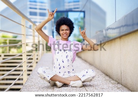 Full body of carefree African American female in casual outfit looking at camera with rock and roll gestures while sitting on street