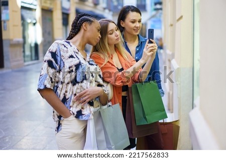 Group of diverse female friends with shopping bags taking picture of showcase and choosing goods in store while standing on street
