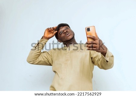 young African man dressed in traditional attire making funny facial expression while taking selfie with phone in hand