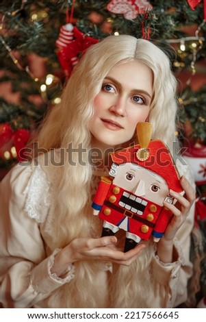 
Portrait of a beautiful white-haired girl with a Nutcracker in her hands against the backdrop of a Christmas tree. New Year. Christmas. Fairy tale for real.