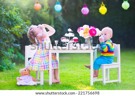 Two happy children, cute curly toddler girl and a little baby boy, brother and sister, enjoying a tea party with their toys playing with dishes, cup cakes and muffins in a sunny summer garden