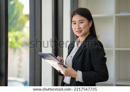 Charming young Asian businesswoman with a smile standing holding a document clipboard at the office. Looking at the camera.
