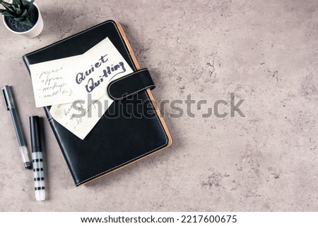 Quiet Quitting. Top view to desk with office supplies. Sticky note with written text Quiet Quitting on closed paper business notebook planner. Quiet quitters perform only required duties of their job. Royalty-Free Stock Photo #2217600675