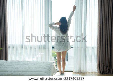 Asian woman standing by the window And stretching in the morning in the bedroom after waking up in bed, fully rested and open the curtains in the morning for fresh air.