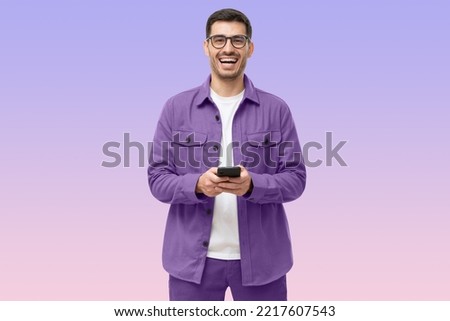 Young happy man laughing as he is looking at camera, holding his phone, isolated on purple background