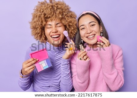 Positive mixed race women apply foundation and colorful palette do makeup want to look beautiful dressed casually have positive expressions isolated over purple background. Cosmetology concept Royalty-Free Stock Photo #2217617875