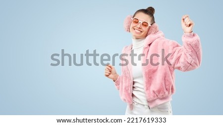 Horizontal banner of young female dancing in pink fluffy bomber, colored glasses and ear muffs, feeling happy at party, isolated on blue background