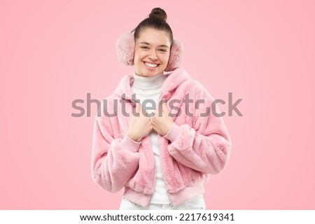 Young smiling girl wearing pink furry jacket and earmuffs, holding hands close to chest, feeling shy but happy, isolated on pink background Royalty-Free Stock Photo #2217619341