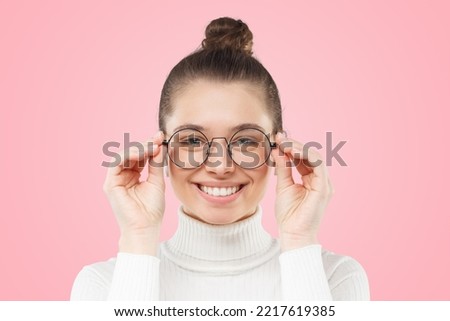 Close-up of smiling young nerdy girl, smart female student touching rim of round glasses as if trying to see something better, isolated on pink background