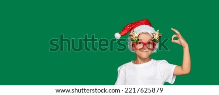 Boy in Santa Claus hat and glasses smiles and shows ok with his fingers. Green background with space for text. Selective focus. Picture for articles about children, holidays, Christmas.