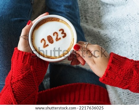 Happy New Year 2023 holidays food art theme, woman in red knitted sweater with jeans holding white coffee cup with number 2023 on frothy surface while relaxed sitting on the couch. (top view) Royalty-Free Stock Photo #2217636025