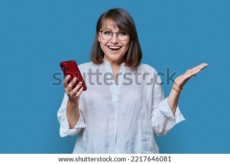 Portrait of middle aged woman with phone looking at camera on blue background