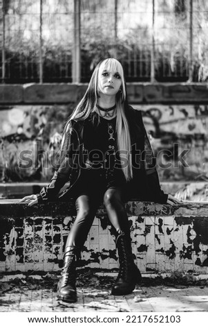 Young and skinny model with long blonde and black hair and black clothes and makeup sitting inside an old abandoned greenhouse (in black and white)