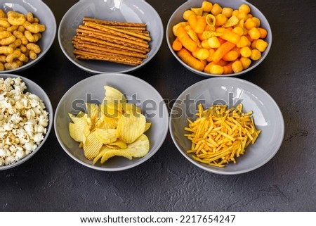 Salty snacks assortment shot on dark slate table. The picture includes potato chips, popcorn, peanut, cheese sticks, ketchup sticks, tzatziki chips, smoky flips, and others