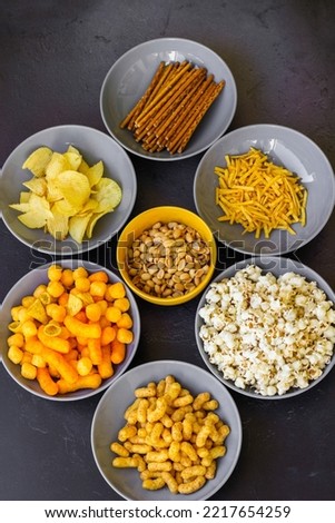 Salty snacks assortment shot on dark slate table. The picture includes potato chips, popcorn, peanut, cheese sticks, ketchup sticks, tzatziki chips, smoky flips, and others