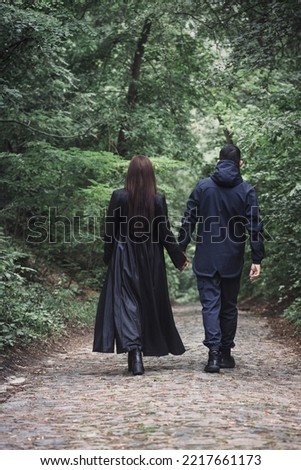 A guy and a girl in black brutal clothes are walking in a dark, foggy forest, back view