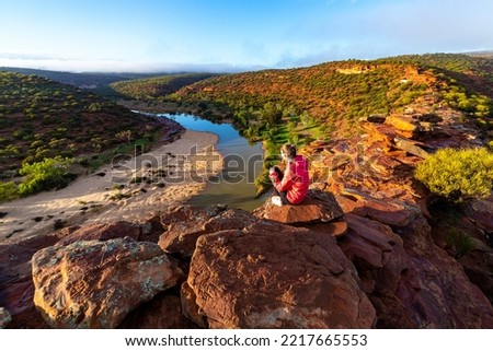 long-haired girl sits on top of a hill on the red rocks of kalbarri national park in western australia; hiking in the wilderness, australian outback Royalty-Free Stock Photo #2217665553