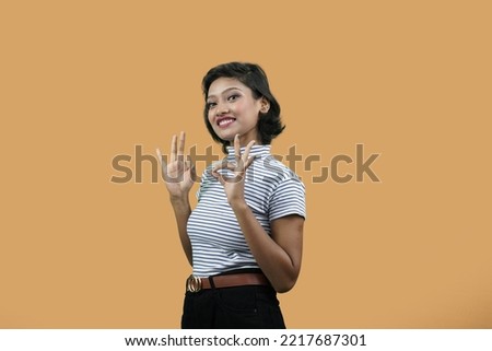 Happy Beautiful Indian girl or Young south Asian woman showing okay gesture with both hands, looking into camera, over orange background.