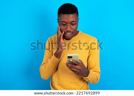 young handsome man wearing yellow sweater over blue background holding in hands cell reading sms using new app 5g