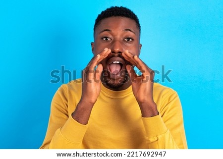 young handsome man wearing yellow sweater over blue background shouting excited to front.