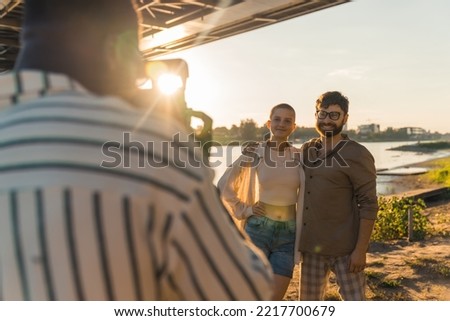 Black blurred male photographer in stripped white shirt taking a photo with his analog camera of his young caucasian friends posing in front of the river under a bridge. Sunshine in the camera. . High