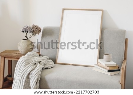 Empty vertical picture frame mockup. Midcentury linen sofa with linen blanket. Ceramic vase with dry hydrangea flowers. Cup of coffee on pile of old books. White wall background. Scandinavian interior