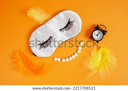 feathers, alarm clock,smile from pills and sleep mask with painted eyelashes on orange background top view insomnia concept
