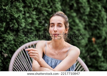 Photo of a girl sitting in a wicker chair on a green terrace, she is dressed in a long loose blue dress. Emotions. Outdoor recreation. Reflections on oneself. Lady in the modern city.