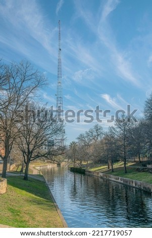 View of a tall signal tower near the River Walk at San Antonio River in Texas. There is a view of a grassy shore with leafless trees and a view of a tower against the cloudy sky background.
