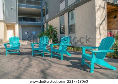 Sky blue wooden lounge arm chairs on a wooden deck outside an apartment building- San Antonio, TX. Four chairs against the view of a building with metal gate below the balcony at the back.