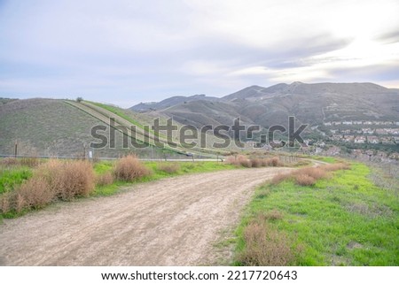 Flattened hiking trail on a mountain with grasses on the side in San Clemente, California. Curved mountain trail with a view of a residential area on the right near the mountain slopes. Royalty-Free Stock Photo #2217720643