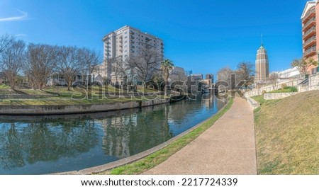 Pathway at River Walk with a view of reflective San Antonio River and residential buildings- Texas. There is a view of a large field with trees near the buildings.