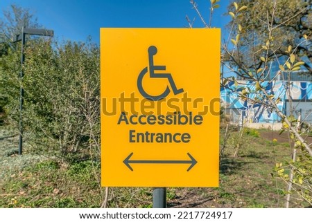 Wheelchair accessible entrance sign with arrow at Waterloo Park in Austin Texas. Bright yellow signage giving information about where handicapped persons should enter the park.