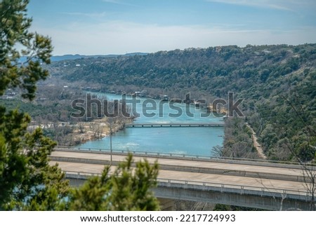 View from Lake Austin Dam of the Colorado River with concrete road bridge. Aerial landscape of water and roadway amid lush foliage of trees and sky on a sunny day. Royalty-Free Stock Photo #2217724993