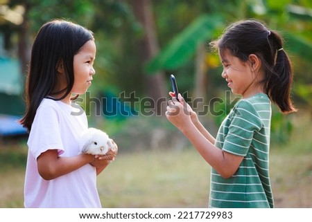 Asian child girl holding a adorable bunny Holland lop on hand and her friend using smartphone taking a picture. They playing with pet together with fun in the yard.