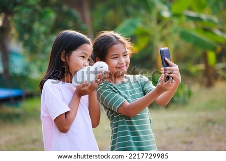 Two asian girls taking a picture and playing with adorable bunny. Sister siblings holding a little rabbit Holland lop on hand and using smartphone selfie together with fun in the garden.