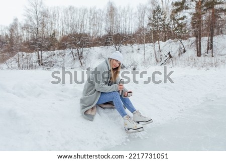 Lovely young woman relaxing after riding ice skates and drinking hot drink from thermo pot on the ice rink. Girl sitting on snow and smiling.