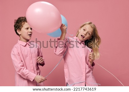 two cute cuddly school age children are standing in pink shirts on a pink background with colorful balloons and smiling happily at the camera. Horizontal studio photography