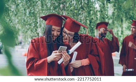 Female graduates are using smartphone looking at screen talking and laughing standing outdoors holding diplomas, pretty girls are wearing formal gowns and hats.